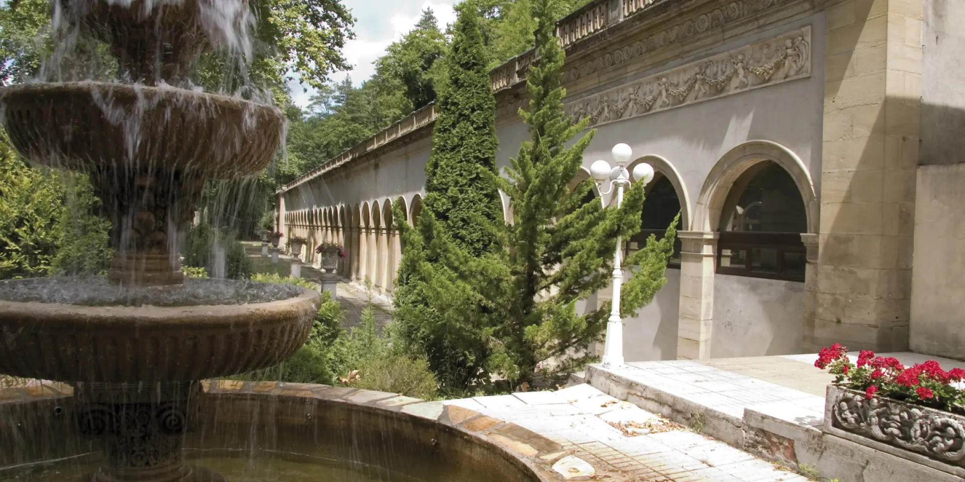 Fuente con varios niveles de agua en un jardín rodeado de vegetación y un edificio antiguo