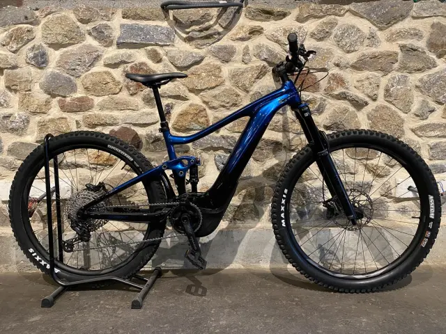 Blue electric mountain bike with black tires and front and rear suspension, leaning against a stone wall